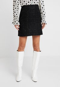 Svart tweed minikjol med texturerat tyg, i rak form. Kombinerad med vita knähöga blanka stövlar och en mönstrad blus.