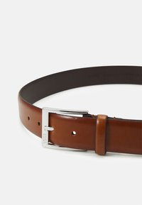 Ceinture en cuir avec un côté intérieur lisse et marron foncé et une surface extérieure brillante, marron clair. Équipée d'une boucle en métal argenté et de plusieurs trous.