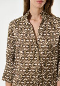 Gemustertes Hemd mit geometrischem Design in Braun-, Creme- und Goldtönen. Es hat einen Kragen, eine Knopfleiste und dreiviertellange Ärmel.