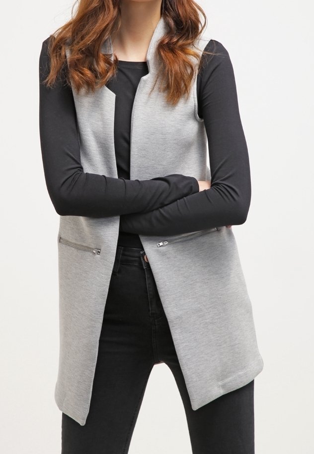Femme aux longs cheveux bruns portant un gilet gris sans manches avec des poches zippées à l'avant sur un haut noir à manches longues et un pantalon noir, bras croisés.