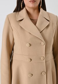 Cappotto beige doppiopetto con finitura testurizzata. Presenta ampie revers, una vita aderente e bottoni circolari dorati come dettagli.