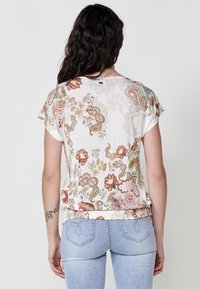 T-shirt à manches courtes à motif floral en tissu crème ; présente des motifs multicolores avec des accents texturés et un ourlet froncé.