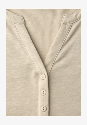 Beiges V-Ausschnitt-Shirt, Vorderseite zeigt Nahtdetails und drei kleine, runde beigefarbene Knöpfe, vertikal in der Mitte angeordnet.