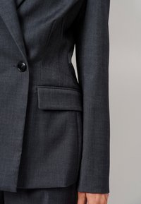 Donkergrijze blazer met een getailleerde pasvorm, enkele knoopsluiting en een zijzak. Beschikt over een gestructureerde stof en een gestructureerde silhouet.