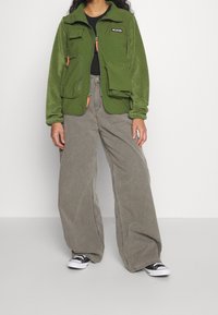 Zelená fleecová bunda s několika kapsami, černé tričko pod tím, v kombinaci s širokými šedými kalhotami a černo-bílými teniskami.