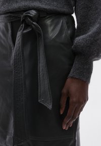 Mustast nahast seelikul on vööl sõlmitud rihm ning õmblustega detailid külgedel; kantud tekstuurse halli kampsuniga.