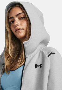 Šviesiai pilka zip-up džemperis su gobtuvu, turintis juodus logotipo akcentus ir mėlyną sportinį marškinėlį po juo. Lygi tekstūra su laisvu siluetu.