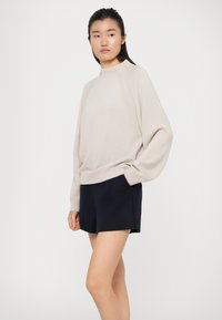 Maglione oversize beige chiaro con colletto girocollo e polsini a coste, abbinato a pantaloni corti blu scuro. Texture morbida, design minimalista.