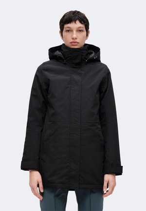Persona con una chaqueta larga de invierno negra con capucha y bolsillos delanteros, de pie frente a un fondo claro liso.