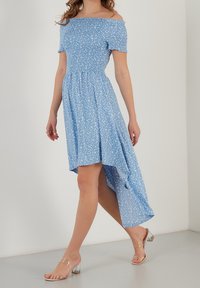 Modré šaty s odkrytémi rameny, s nařaseným korzetem, květinovým vzorem a asymetrickým lemem, doplněné průhlednými sandály na blokovém podpatku.