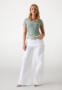 T-shirt fitted verde menta con la scritta "Guess" in lettera d'argento, abbinato a pantaloni bianchi a vita alta e gamba larga e una cintura d'argento texturizzata.