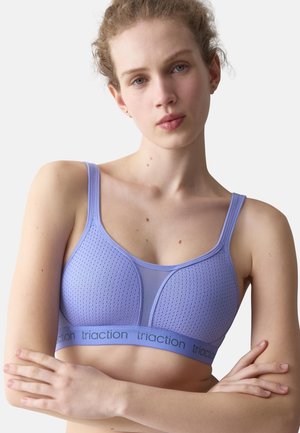 Jeune femme à la peau claire et aux cheveux bouclés porte un soutien-gorge de sport violet perforé avec le logo "triaction", bras croisés sur un fond uni.