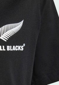 Czarny bawełniany t-shirt z białym graficznym wzorem paproci i napisem "ALL BLACKS". Krótkie rękawy i okrągły dekolt z gładką fakturą.