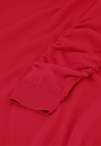 Roter Pullover mit langen Ärmeln und einem gerafften, elastischen Manschettendetail an einem Ärmel, das Textur und Stofffalten zeigt.