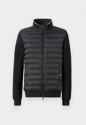 Sort quiltet jakke med fuld lynlås, ståkrave med rød og hvid kant, lange ærmer og sidelommer med trykknapper.