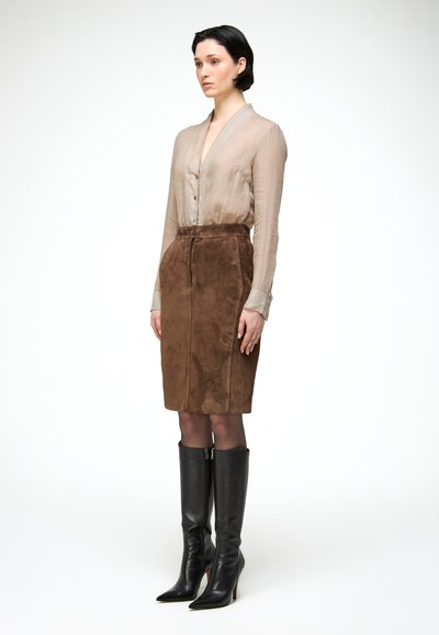 Femme debout, portant un chemisier beige transparent, une jupe en daim marron longueur genou, des collants noirs et des bottes noires à talons montant jusqu'aux genoux.