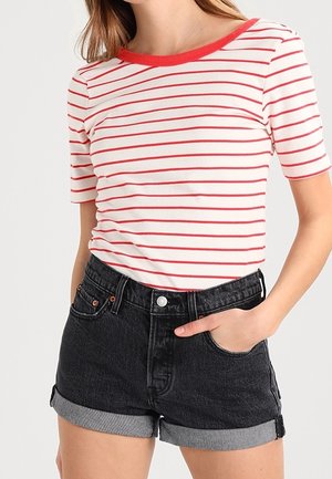 Kobieta w białej koszuli z czerwonymi poziomymi paskami i czerwonym dekoltem, w połączeniu z podwiniętymi czarnymi szortami dżinsowymi, ręka w kieszeni.