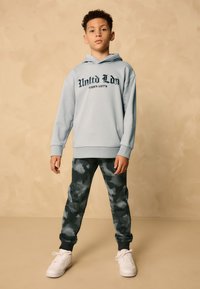 Felpa con cappuccio azzurro chiaro con scritte nere grafiche, abbinata a pantaloni jogger con motivo mimetico scuro. Completa il look un paio di sneakers bianche.