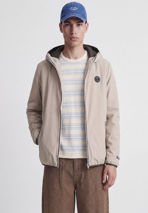 Jeune homme portant une veste à capuche beige zippée, une chemise rayée bleu clair et beige, un pantalon marron et une casquette bleue avec le texte "utility studios".