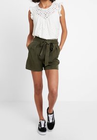 Haut blanc en broderie anglaise avec manches à volants, associé à un short taille haute vert olive doté d'une ceinture nouée. Des baskets noires complètent le look.