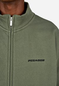 Tuvumā ir tumšādains cilvēks, kurš valkā olīvu zaļa krāsas jaku ar rāvējslēdzēju, uz krūtīm ir izšūts vārds "PEGADOR".