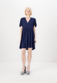 Abito blu navy con scollatura a V e maniche corte a volant. Tessuto fluido con vita arricciata e orlo posteriore leggermente più lungo. Un paio di scarpe con zeppa grigie.