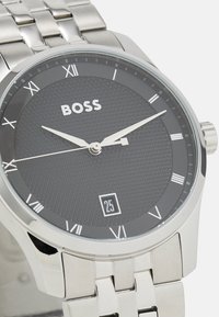 Montre en acier inoxydable argenté avec un cadran rond en gris texturé, des chiffres romains et un affichage de la date à la position 6 heures.