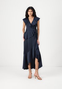Vestido azul-marinho com decote em V, detalhes de babados, bainha assimétrica e uma silhueta fluida, combinado com sandálias de salto alto bege com tiras.