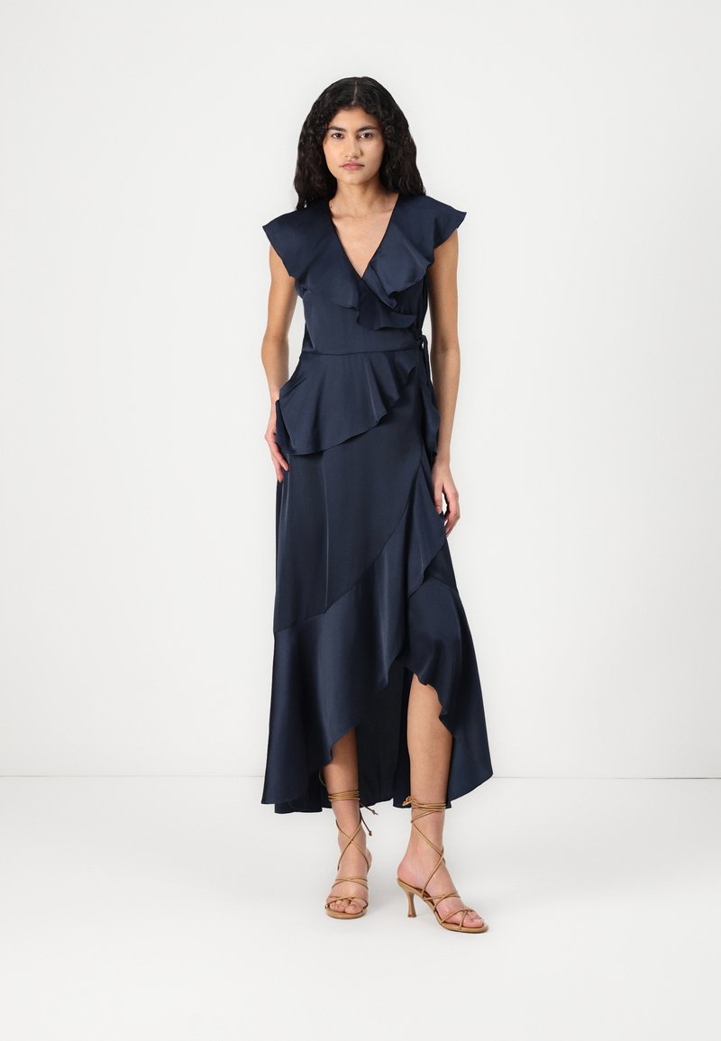 Vestido azul-marinho com decote em V, detalhes de babados, bainha assimétrica e uma silhueta fluida, combinado com sandálias de salto alto bege com tiras.