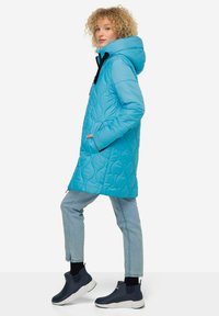 Padded coat in bright blue with a hood, featuring a quilted pattern, two side pockets, and a black zipper. Paired with light blue jeans and dark boots.