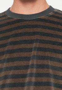 Λήψη κοντινής εικόνας ενός ατόμου που φοράει ένα καφέ και μαύρο, οριζόντια ριγέ μπλουζάκι με λαιμόκοψη.