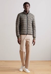 Padded olive green jacket with vertical stitching, zip closure, and ribbed cuffs, paired with light beige trousers and white sneakers.