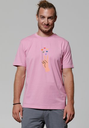Roze katoenen T-shirt met een afbeelding van een hand die een vredesteken maakt, met kleurrijke harten erboven. Korte mouwen en een ronde hals.