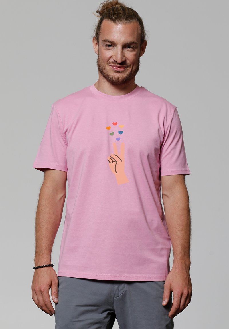 T-shirt en coton rose avec un graphique d'une main faisant un signe de paix, accompagnée de cœurs colorés au-dessus. Manches courtes et col rond.