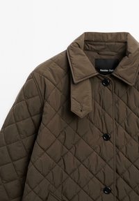Gequilte bruine buitenkleding met een kraag, voorzien van een knoopsluiting aan de voorkant, zijzakken en een getextureerd ruitpatroon. Tag zichtbaar binnenin de kraag.