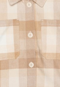 Beige geruit overhemd van zachte stof, met een puntige kraag, twee borstzakken en crèmekleurige knopen, met verschillende ruitpatronen.