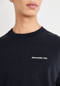 Μαύρο βαμβακερό t-shirt με στρογγυλή λαιμόκοψη, με λευκό κεντητό λογότυπο "Abercrombie | NYC" στο στήθος. Λείος υφασμάτινος χαρακτήρας, άνετη εφαρμογή.