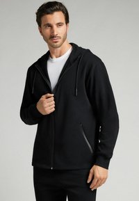 Sudadera con cremallera de color negro, fabricada en tejido suave, con capucha ajustable, dos bolsillos laterales con cremalleras y un corte relajado.