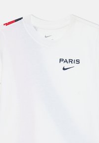 White cotton T-shirt featuring "PARIS" and Nike logo in navy embroidery. Left shoulder has a color-block design with red and blue accents.