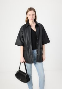 Geacă oversized din piele neagră, cu mâneci scurte, nasturi pe față și două buzunare laterale, purtată cu blugi albaștri deschiși și o geantă de mână neagră.