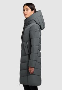 Femme portant un long manteau rembourré gris avec un col haut, une grande capuche et une poche latérale zippée, se tenant de profil contre un fond uni.