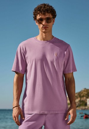 Junger Mann trägt ein lila T-Shirt und Shorts mit brauner Sonnenbrille und steht am Meer unter klarem blauem Himmel.