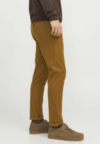 Chinos en coton marron avec une coupe slim, ourlet à revers et poches latérales, associés à des chaussures lacées gris-marron et des chaussettes marron.