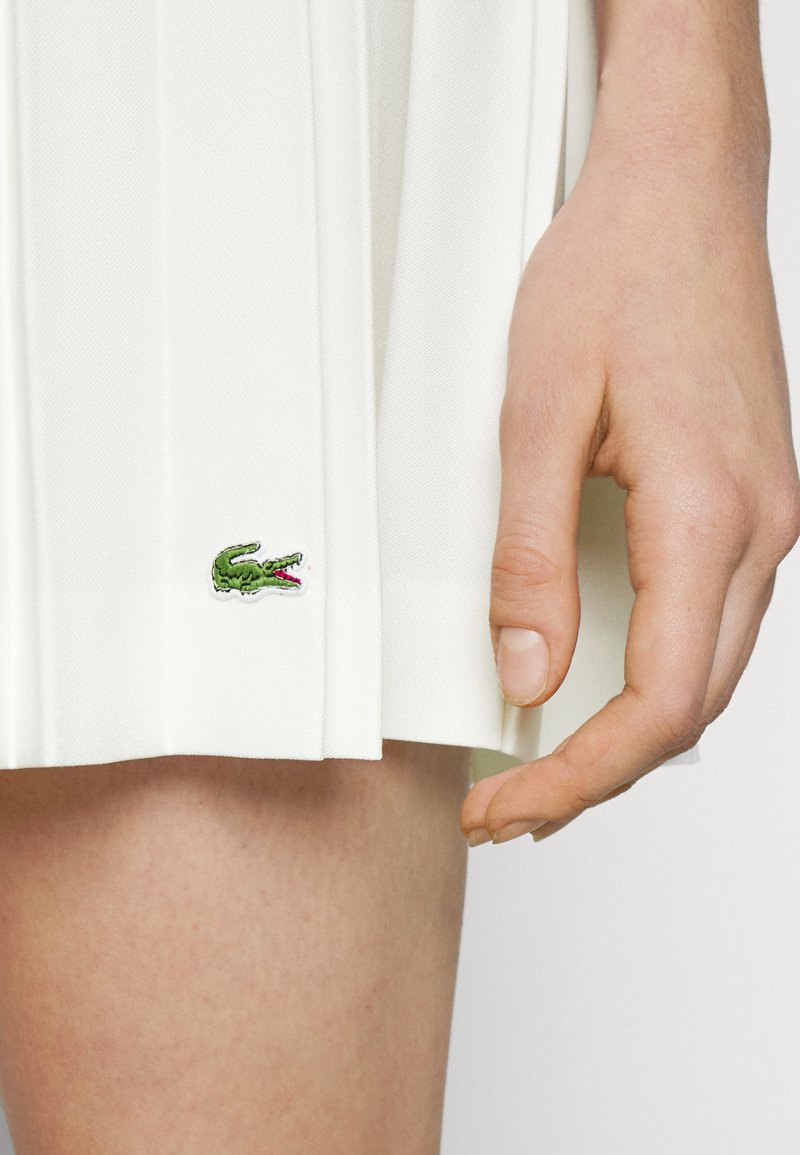 Weiße plissierte Skirt aus strukturiertem Stoff. Hat ein kleines, grün gesticktes Krokodil-Logo am Saum. Hand, die das Kleidungsstück berührt.