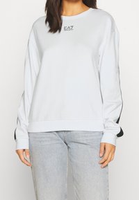 Donna che indossa un sweatshirt bianco a maniche lunghe con dettagli a strisce nere e logo EA7 Emporio Armani, abbinato a jeans blu chiaro.