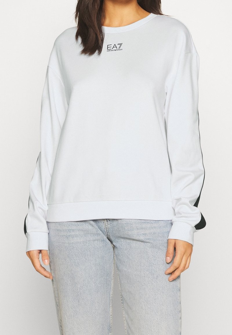 Donna che indossa un sweatshirt bianco a maniche lunghe con dettagli a strisce nere e logo EA7 Emporio Armani, abbinato a jeans blu chiaro.