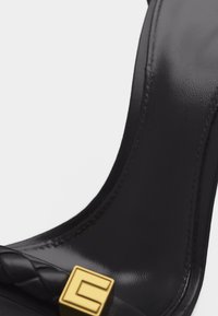 Κοντινό πλάνο ενός μαύρου δερμάτινου λουριού γοβών με σχέδιο πλεξούδας και χρυσή τετράγωνη αγκράφα ως διακοσμητικό στοιχείο.