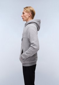 Felpa grigia con cappuccio, dotata di tasca frontale, cordino e polsini a costine. Indossata sopra pantaloni neri, vista di lato. Tessuto liscio.