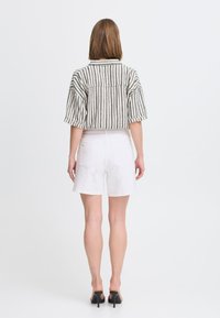 Camisa de manga corta a rayas en beige y marino, con un cuello y un detalle de bolsillo en la parte posterior, combinada con pantalones cortos de denim blancos. Tacones negros destalonados.