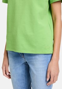 Zielona, teksturowana koszulka z krótkim rękawem, z prążkowanym wzorem i detalem logo na dolnym końcu. W połączeniu z jasnoniebieskimi dżinsami.